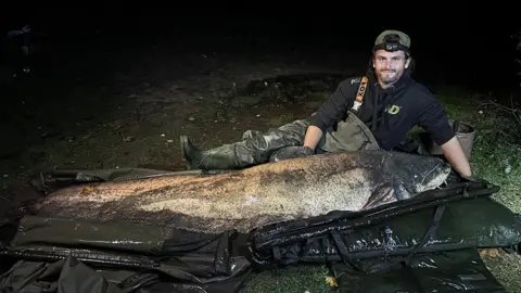 Chloe Ing Shaun Ing, lying on the ground next to a large catfish in a night time image taken using a flash.