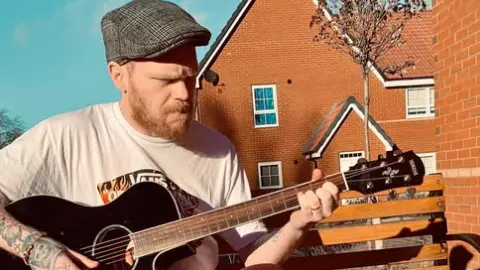 Craig Brown Craig Brown, wearing a flat cap and t-shirt, sits on a bench outside while playing guitar.