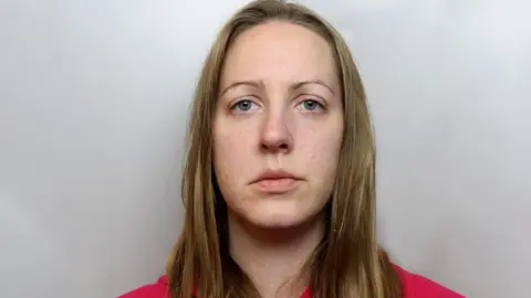 Cheshire Constabulary Mugshot of Lucy Letby wearing a red hoody. she has blonde hair and thin eyebrows.