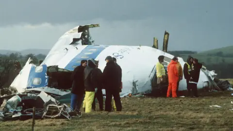 Reuters Emergency workers survey some of the wreckage of Pan Am Flight 103. There are several people standing around some of the wreckage of the plane.