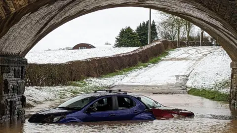 Supplied Two cars - one blue and one red - stuck in flooding under a bridge. Snowy fields and a road can be seen in the background.