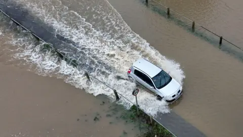 PA A car driving through a flooded road with water splashing around, partially submerging the road surface, and a 40 mph speed limit sign visible nearby.