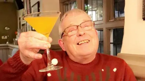 Family Photo A grey-haired man with glasses smiling and holding up a yellow cocktail