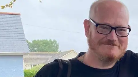 Met Police Ian Airlie is seen with glasses and a beard is smiling outdoors, with houses and trees visible in the background on a cloudy day.