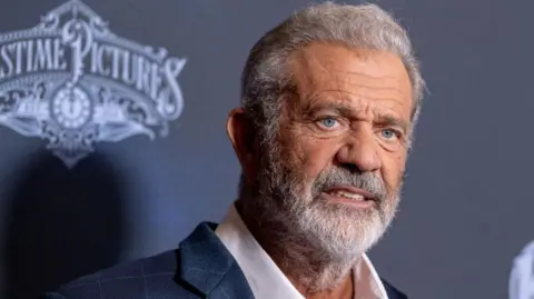 Getty Images Mel Gibson, wearing a dark blazer and white shirt with an open collar, with grey hair and a grey beard, poses for photographs at a screening in Los Angeles