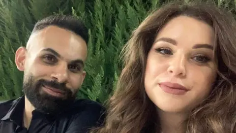 BBC Ashraf and Julia both smiling for the camera, with a background of greenery. Ashraf has dark hair and a beard and is wearing a dark shirt, and Julia has lighter brown hair - long and wavy - and has brightly painted lips.
