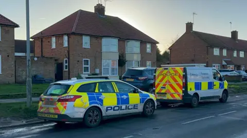 Ben Parker/BBC A police car and police forensic van are parked at the side of Hawthorn Drive. Houses can be seen to the left of them with other vehicles parked on a grass verge.