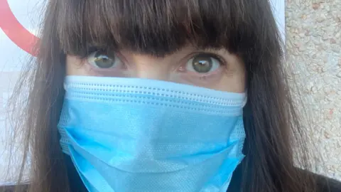 BBC A close up selfie of journalist Nicola Bryan's face. She is wearing a blue face mask and has long brown hair.