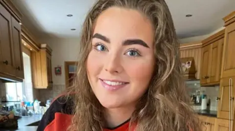 Family Handout Jeni Larmour pictures in her kitchen, smiling wearing a red t-shirt.