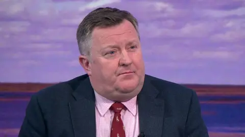 BBC Prof Taylor is wearing a navy blazer with a pinkish shirt. He has a red tie on. 