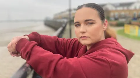 BBC/Piers Hopkirk Danielle wearing a red hoodie looking to camera leaning over a railings on an unidentified seafront