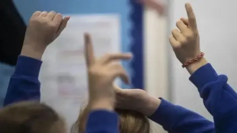 PA Media Children's hands are pointed in the air as if to ask a question, they are wearing blue jumpers and one has a bracelet.