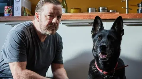 Netflix Ricky Gervais is on the left in a grey T-shirt looking at the camera. A German shepherd dog is on the right and cupboards are behind them.