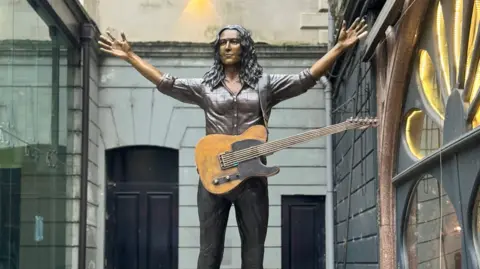 BBC Bronze statue of Rory Gallagher with a guitar hanging off him, photographed against brick wall at Ulster Hall