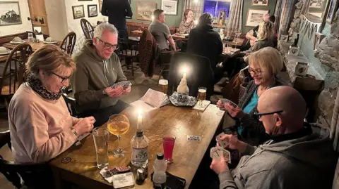Tan Hill Inn Pub visitors sit around a candle-lit table enjoying drinks and playing cards.