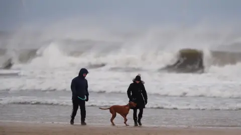 PA Media Dog walkers in the wind on Tynemouth Longsands on the North East coast of England. Large waves have formed in the background.