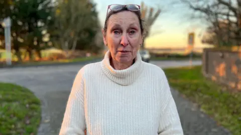BBC A woman in a white jumper stand by a road outside her house.