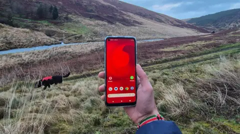Vodafone A man holds a mobile phone in the middle of a mountain in West Wales, tere is green space and blue sky, with a river and a dog.
