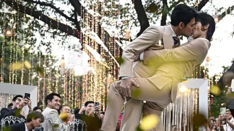 Getty Images This photo taken on January 10, 2025 shows Thai actors Apiwat "Porsch" Apiwatsayree (L) and Sappanyoo "Arm" Panatkool in beige tuxedos sharing a kiss at their unofficial wedding ceremony in Bangkok. They are surrounded by family and friends taking photos of them under a tree decorated with twinkling lights.