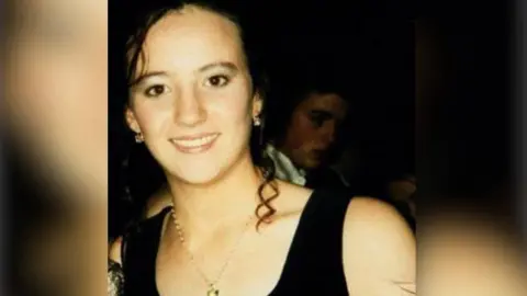 Marlow family A young woman wears gold earrings and necklace. She has dark brown hair and brown eyes. She smiles at the camera, white teeth showing. Behind her is the face of a boy half hidden by shadow. He wears a white shirt.