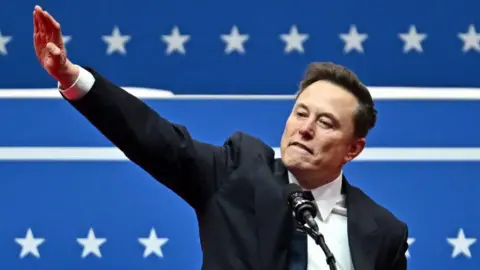 Getty Images Elon Musk at a rally in front of a microphone. His right arm is raised out at a 45 degree angle to his body, with his palm flat and pointing downwards.