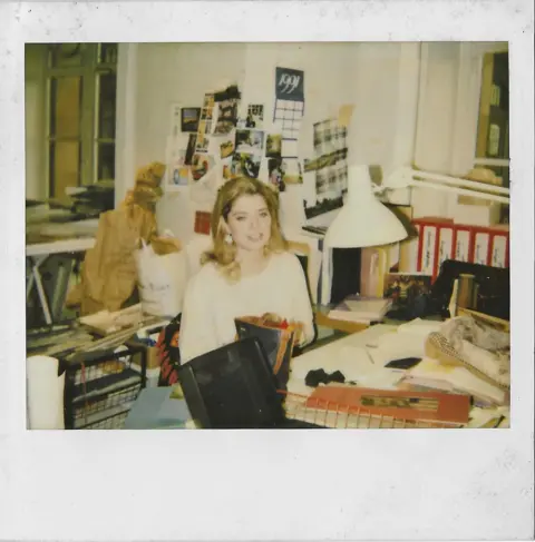 Frances A polaroid picture taken in 1991 of Frances inside the Harrods office. She is smiling and sitting behind a desk busy with papers and books. Behind her are red files and a notice board with a calendar, pictures and more papers attached.
