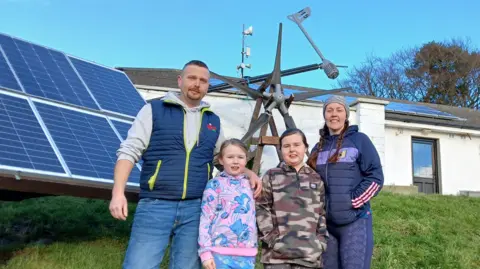Michael Wilkinson Michael Wilkinson with his wife and two daughters, standing outside their home in County Kilkenny. Two dogs are in the background in their tiered garden. There are solar panels on the roof and propped up on the lawn. There is also a wind turbine behind them.