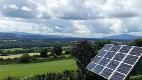 Michael Wilkinson A board of solar panels set on a sloped garden outside the Wilkinson family's home on a sunny day in County Kilkenny. The board can be swivelled to point the panels at the sun's rays. The garden is surrounded by the green fields of the Suir Valley with hills in the distance.