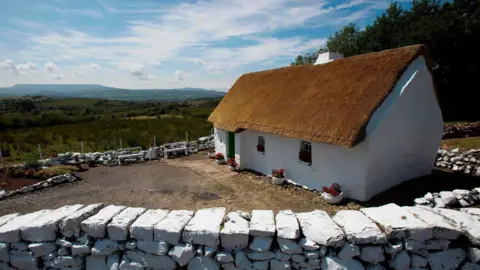 PAUL FAITH/AFP via Getty Images Mullylusty Cottage, a thatched home outside Belcoo in County Fermanagh. The small three-room building is white washed and has three windows and a door at the front. There is a dry stone wall around the house, which overlooks fields and distant hills.