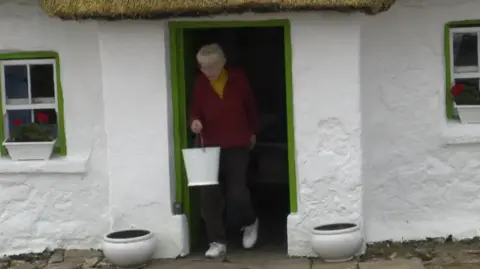 A screen grab of Margaret Gallagher when she was filmed by the BBC in 2019 leaving her cottage to fill a bucket of water from her well. She is wearing a red cardigan over a yellow top, dark trousers and white trainers.