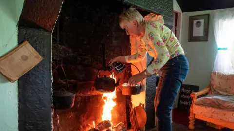 PAUL FAITH/AFP via Getty Images Margaret Gallagher heating a kettle of water at her large open fireplace in Mullylusty Cottage near Belcoo in 2019. She has short, grey hair and glasses and is wearing a yellow floral T-shirt and blue jeans.