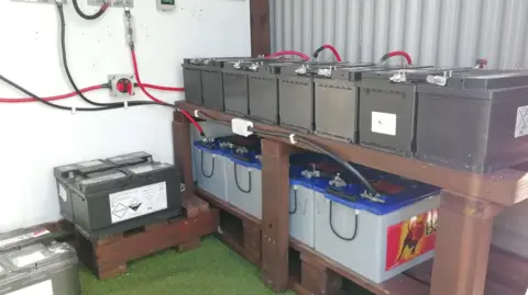Michael Wilkinson A battery bank at the Wilkinson's home in County Kilkenny, Several large batteries are stacked on wooden shelving. They are connected to power sources with cables.