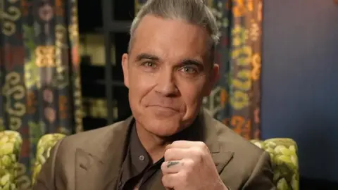 Tony Dolce/BBC Robbie Williams pictured smiling whilst raising a fist to the camera. He has short greying hair, pulled back from his forehead, and his wearing a light brown suit over a dark brown collarless shirt.