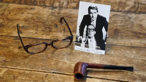 PA Media Eric Morecambe's glasses and pipe resting on wooden planks. The glasses are brown imitation tortoiseshell and their arms are open. The pipe is lying on its side and is brown. There is also a black and white photo showing Eric Morecambe with Ernie Wise.