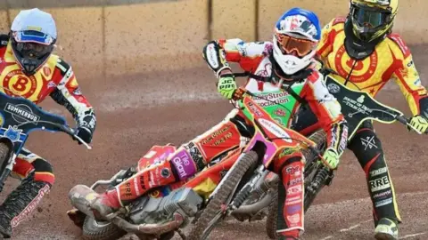 Three speedway riders battle it out on the track. They are wearing multi-coloured race suits