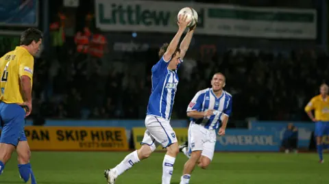 Paul Hazlewood/BHAFC Jake in 2006 running and holding the ball above his head after scoring three goals. He is wearing a blue and white striped kit with white shorts and socks. The opposition are wearing yellow and walking away and there is a small stand of the Withdean stadium behind him with yellow and blue advertising hoardings between the pitch and stand