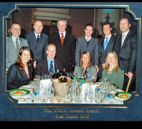 Picture of Rachel Reeves in a group of smiling at the camera. Reeves is sitting next to a man, and there are two other women seated at a table, on which there are bottles and wine glasses. Six men stand behind the seated group in a semi circle.
