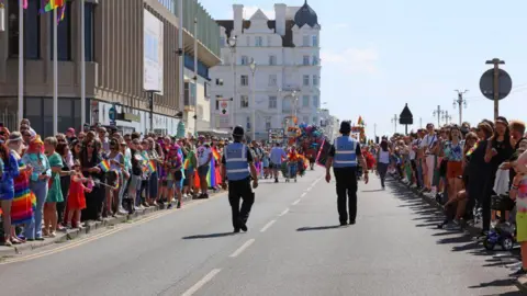 Getty Images People line the streets of Brighton for the Brighton Pride parade. There are two police officers in the middle of the road, and hundreds either side of it.