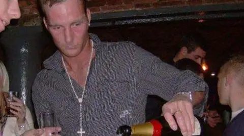 UGC Adam Minett pours a bottle of champagne with his left hand. He is wearing an open-necked shirt with a chain around his neck, and is in the middle of a crowd in a bar in Liverpool.