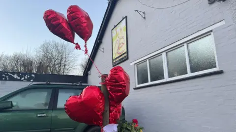 BBC/JO BURN Four heart shaped balloons are tethered to a gatepost outside the Three Horseshoes pub in Knockholt. A green car is parked behind the balloons.