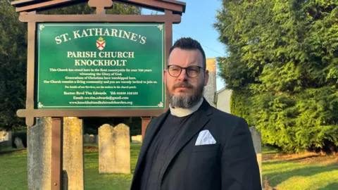 Rev Tim Edwards stands in front of the church sign for St Katherine's in Knockholt. He wears clerical dress and has a beard and glasses, with a handkerchief in his top pocket.