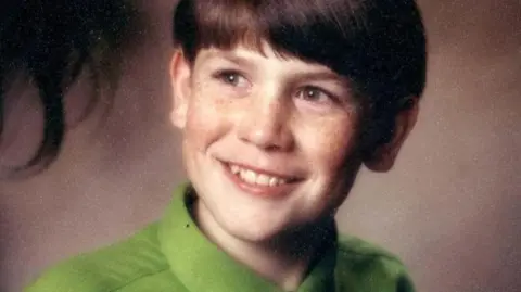 Barker family A young boy with brown hair smiles at something off-camera. He is wearing a bright green shirt. 