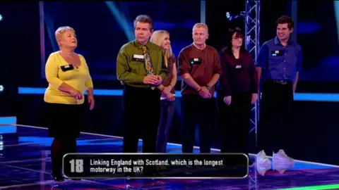 Breakaway/Gogglebox Cornelius wearing a green shirt and brightly coloured tie standing with other contestants on Breakaway in 2012, with a question on the screen that says: Linking England with Scotland, which is the longest motorway in the UK"?