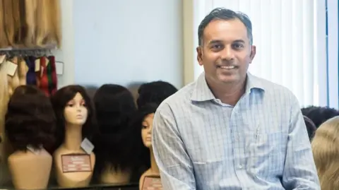George Cherion George Cherion smiling sits in front of mannequins wearing wigs.