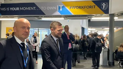 Three men in suits and ties standing in a busy area in a school with lots of school pupils passing