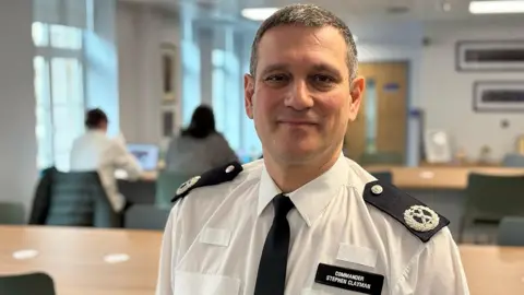 Commander Stephen Clayman in his police uniform with name badge, in an office, two colleagues visible in the background.