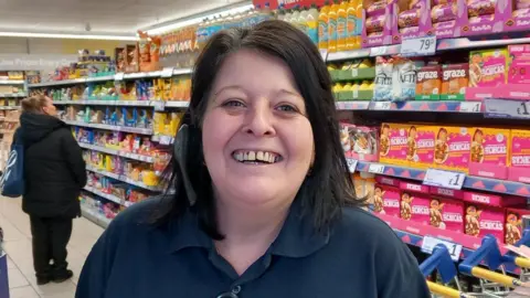 A woman with black hair and wearing a headset is smiling at the camera in a supermarket.