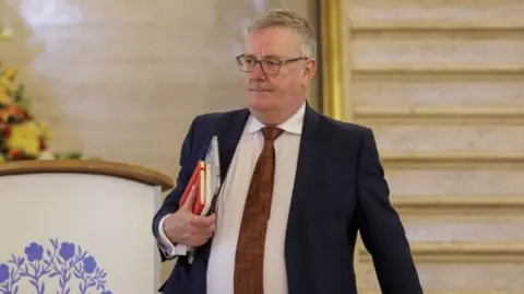 PA Media Mike Nesbitt walking out of the Northern Ireland Assembly with files under on arm. He has salt and pepper hair and black-framed glasses and is wearing a dark suit jacket, white shirt and red tie.
