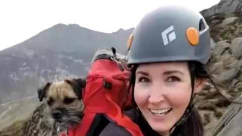 Aly Coyle Aly Coyle wearing a helmet, she had long brown hair tied up, she's carrying a red backpack with her dog inside (a terrier type dog with light brown and grey hair). Aly is climbing Cavehill smiling at the camera.