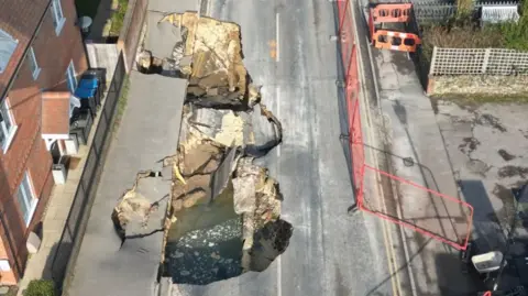 Eddie Mitchell A large sinkhole in Godstone High Street. There is a red brick house on the left and red fencing on the right of the street.
